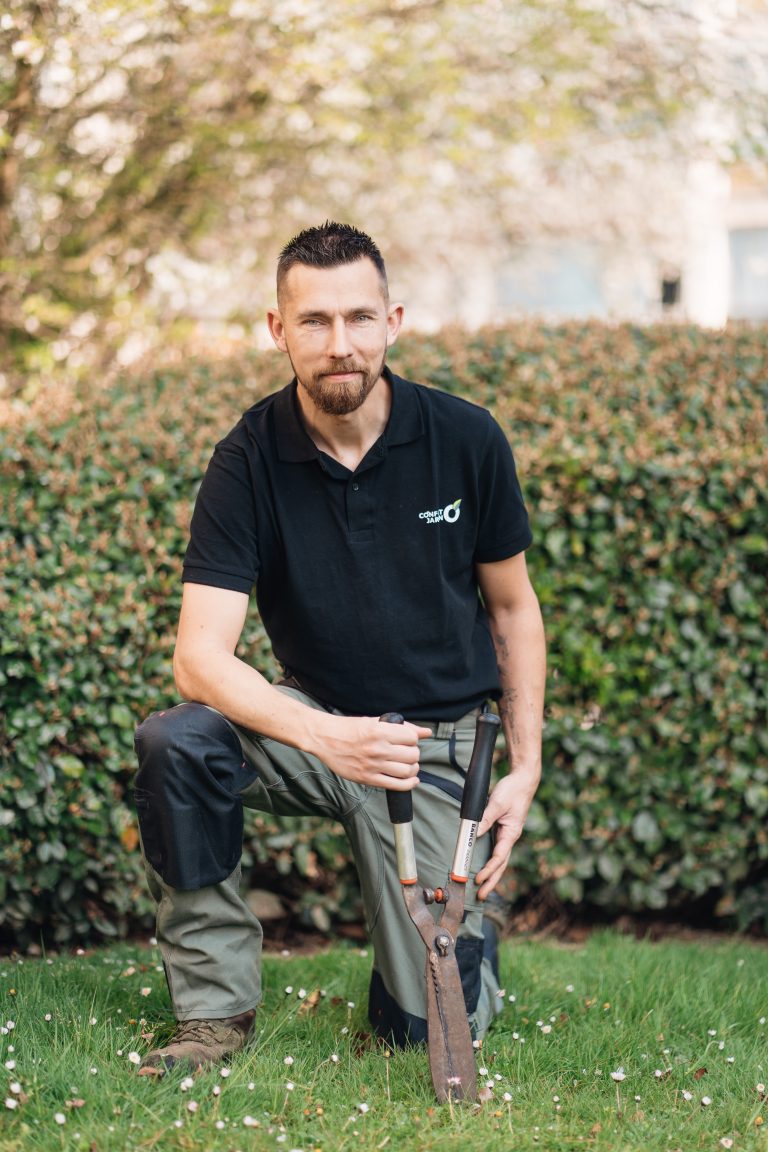 Stéphane, paysagiste CONFORT O JARDIN, un genou à terre devant une haie, coupe-branches à la main