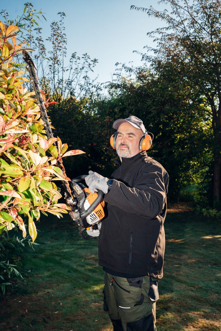 Renaud taille une haie de photinias au taille-haie thermique, casque antibruit sur les oreilles