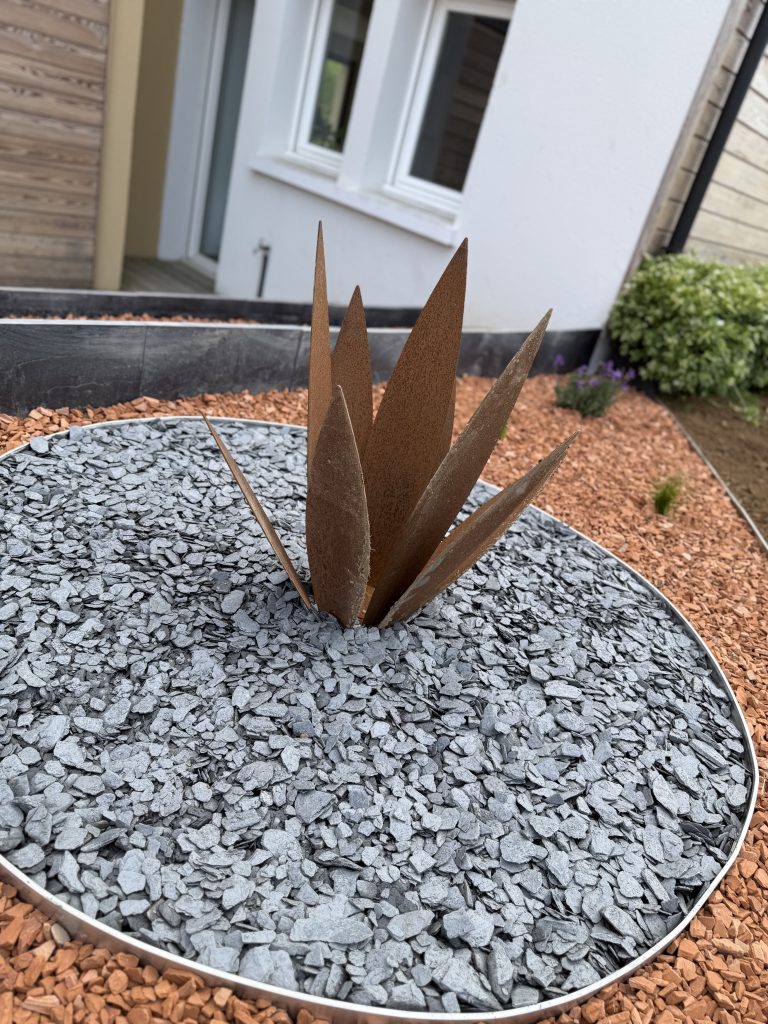 Îlot de paillage d'ardoise cerclé d'aluminium avec sculpture végétale en acier corten, au milieu de la pouzzolane