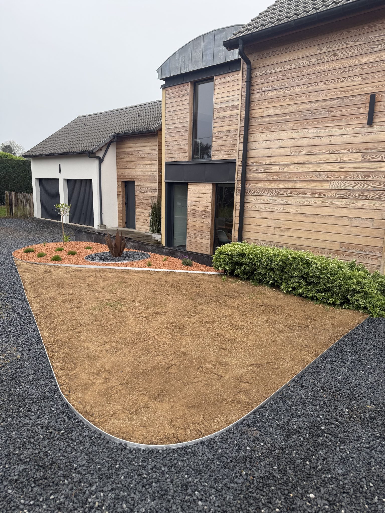 Pelouse en forme bordée d'aluminium et entourée de gravier noir, devant une maison en bardage bois, aménagée par CONFORT O JARDIN